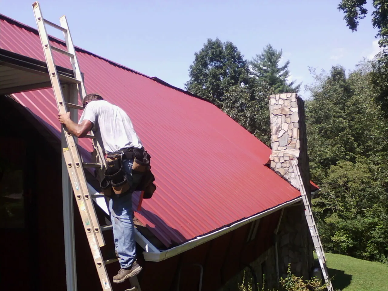 Roofer on a ladder beside a red metal roof and stone chimney — Soffit & Fascia Repair by Roofing Brothers in Great Lakes
